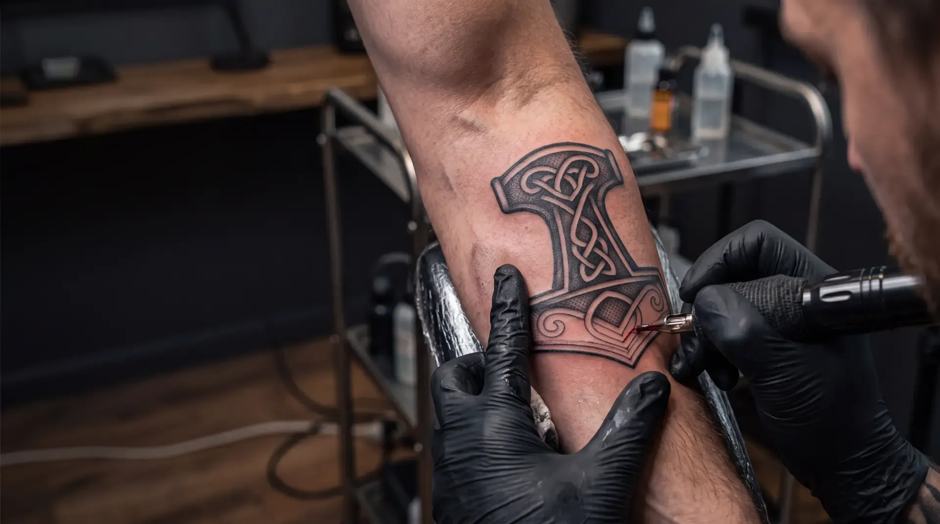 A tattoo artist inking a Mjölnir (Thor's hammer) tattoo with Norse knotwork on a forearm
